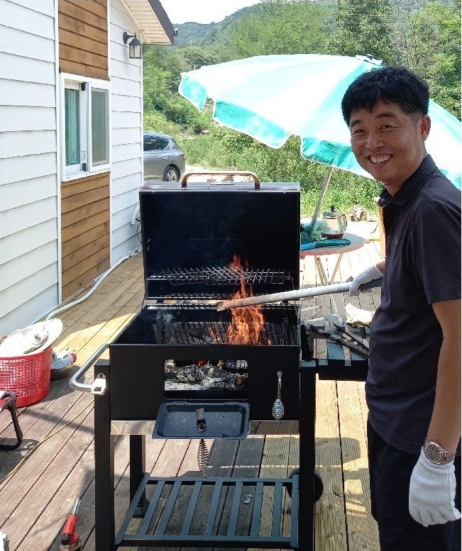 펜션 데크의 바베큐 그릴 앞에서 불을 살피며 웃고 있는 농장주